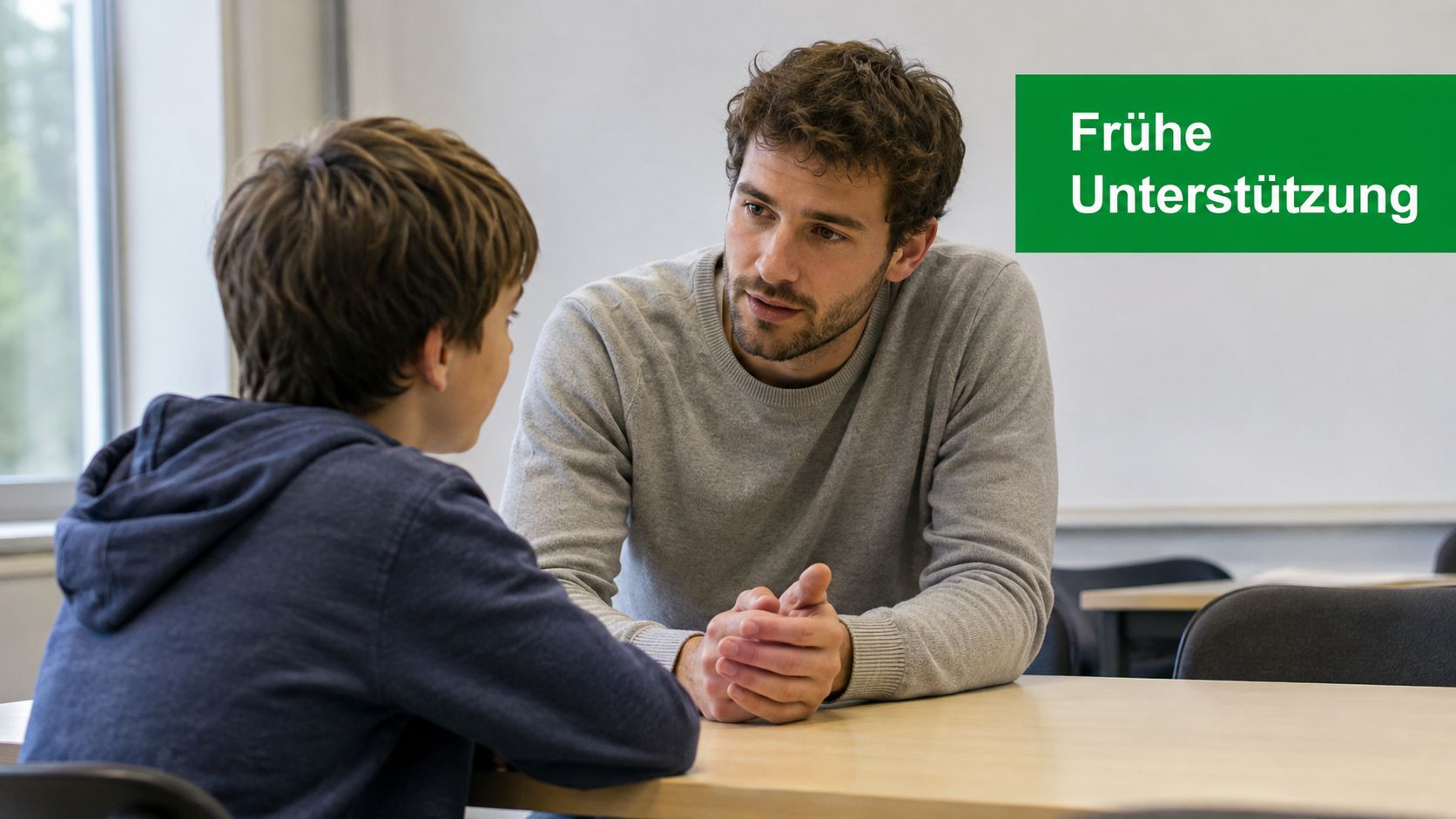 Ein Sozialpädagoge spricht unterstützend und einfühlsam mit einem jungen Schüler an einem Tisch im Klassenzimmer.